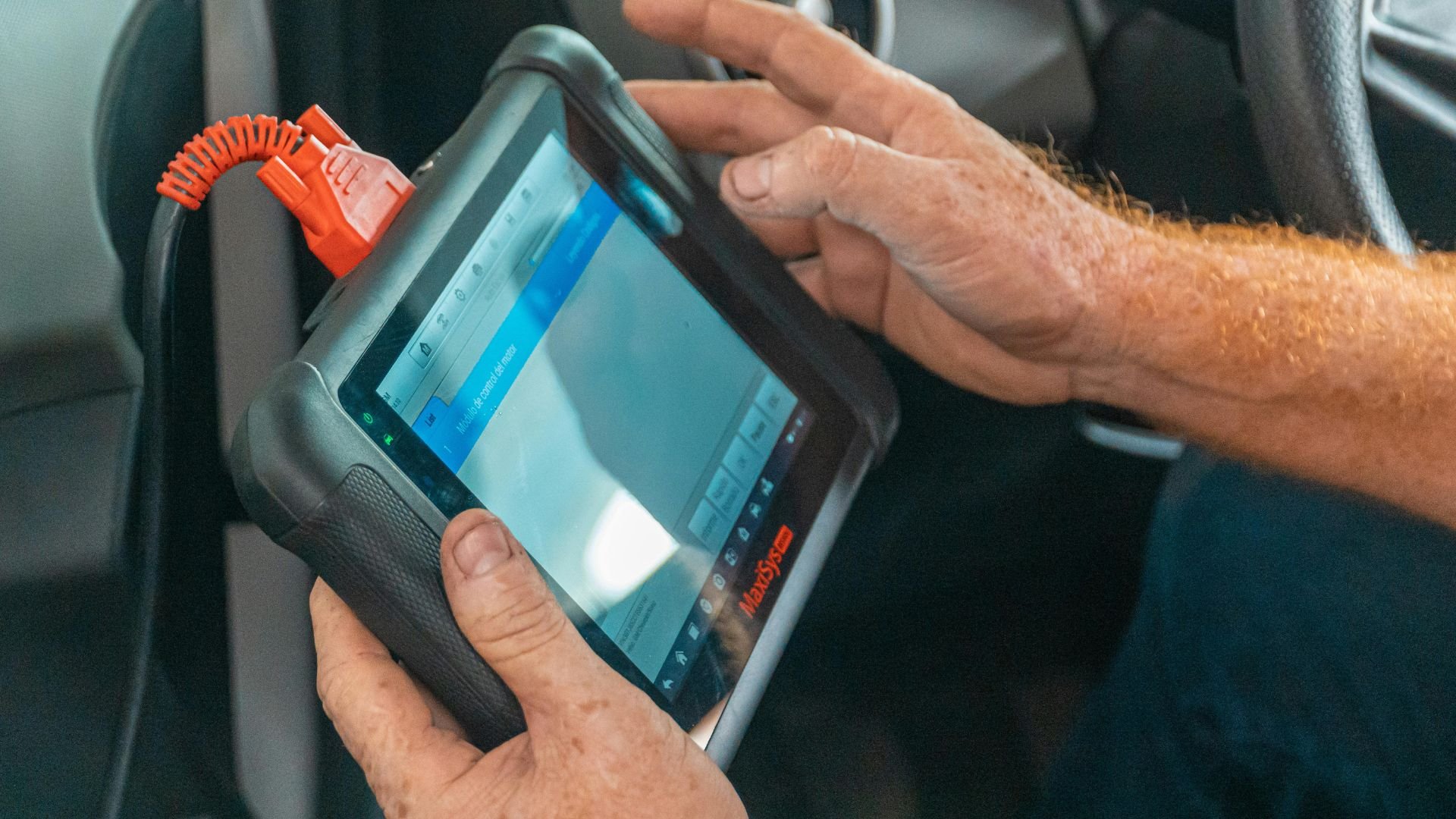 Hands holding diagnostic tablet with orange connector in vehicle interior